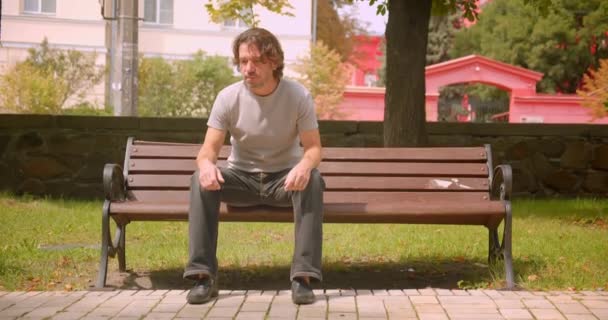 Closeup portréja csinos kaukázusi ember ül a padon elgondolkodva a parkban a szabadban — Stock videók