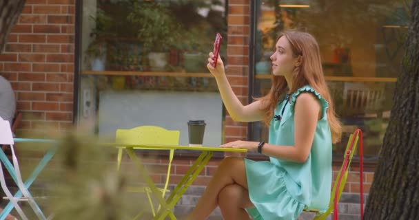 Gros plan portrait de jeune belle femme caucasienne navigation sur le téléphone et boire du café assis dans le café à l'extérieur — Video