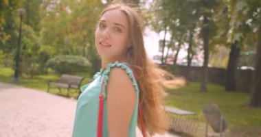 Yakın çekim geri görünüm portre genç sevimli kadın yürüyüş dönüm ve parkta mutlu gülümseyen gurur saç ile kameraya bakarak