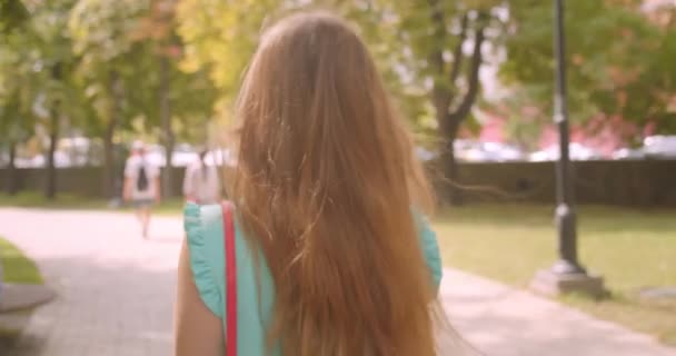 Nahaufnahme Rückseite Porträt einer jungen hübschen Frau, die sich dreht und mit schmeichelhaftem Haar glücklich lächelnd in den Park blickt — Stockvideo