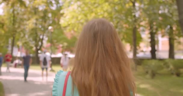 Gros plan vue de dos portrait de jeune belle femme marchant tournant et regardant la caméra avec des cheveux flatteurs souriant joyeusement dans le parc — Video