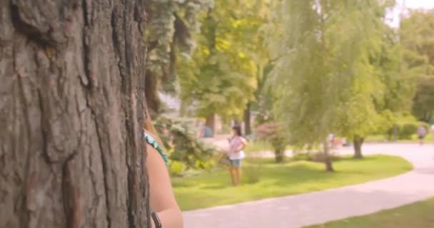 Primer planoretrato de joven de pelo largo hermosa hembra escondida detrás del árbol mirando a la cámara con pelo halagador en el parque al aire libre — Vídeo de stock