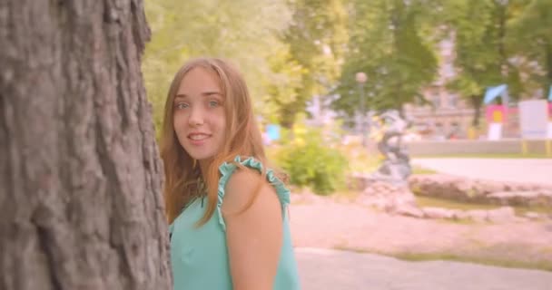 若いかわいい長い髪の美しい女性のクローズアップポートレートは、屋外の公園でお世辞の髪でカメラを見て木の後ろに隠れています — ストック動画