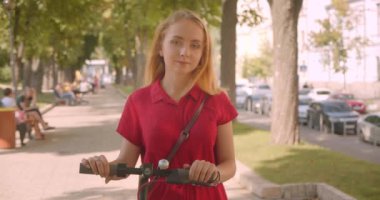 Park açık havada duran kameraya bakan tekme scooter tutan kırmızı elbiseli genç güzel beyaz beyaz kadın Closeup portre