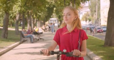 Kırmızı elbiseli genç güzel beyaz kadın kick scooter tutan closeup portre kamera neşeyle park açık havada ayakta gülümseyerek bakarak