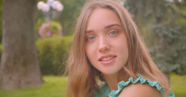 Closeup retrato de jovem de cabelos longos bela fêmea sentada na grama olhando para a câmera descansando alegremente no parque ao ar livre — Vídeo de Stock