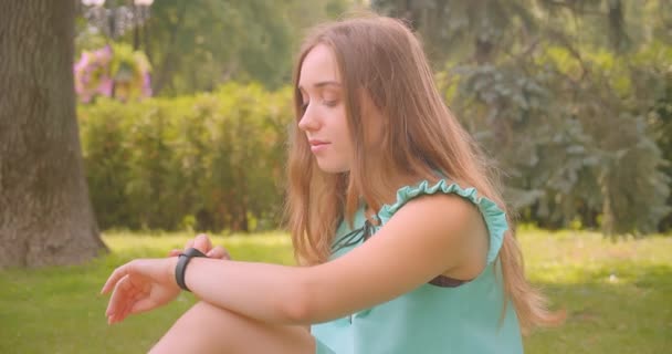 屋外の公園で休んでいるスマートウォッチを使用して草の上に座っている若い長い髪の美しい女性のクローズアップ肖像画 — ストック動画