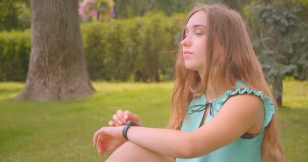 Primo piano ritratto di giovane carino dai capelli lunghi bella femmina seduta su erba verde con orologio intelligente nel parco all'aperto — Video Stock