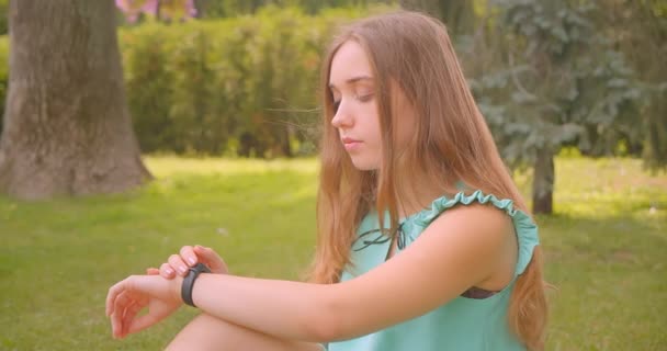 Primer plano retrato de joven lindo pelo largo hermosa hembra sentada en la hierba usando reloj inteligente en el parque al aire libre — Vídeos de Stock