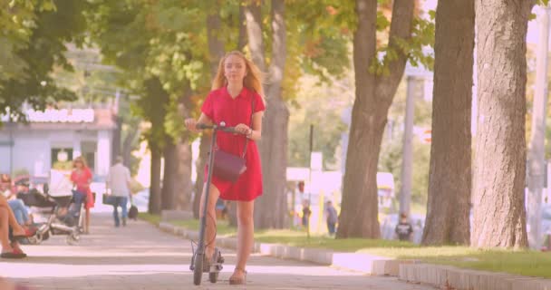 Retrato de joven hermosa mujer caucásica en vestido rojo sosteniendo patada scooter mirando a la cámara de pie en el parque al aire libre — Vídeos de Stock