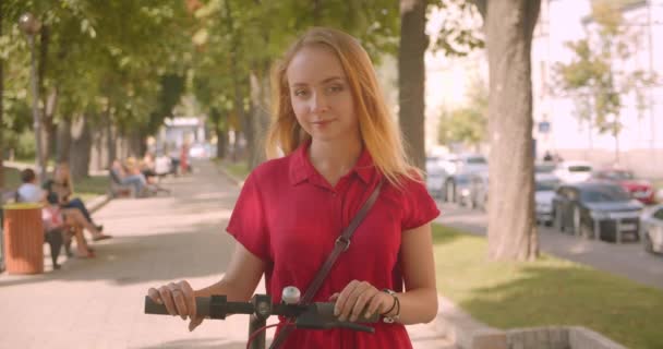 Nahaufnahme Porträt einer jungen schönen kaukasischen Frau im roten Kleid, die einen Tretroller in die Kamera hält und glücklich lächelnd im Park im Freien steht — Stockvideo