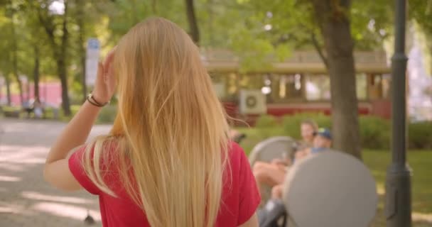 Primer plano vista trasera retrato de la joven rubia de pelo largo linda hembra caminando en el parque girando y mirando a la cámara con su pelo halagador al aire libre — Vídeos de Stock