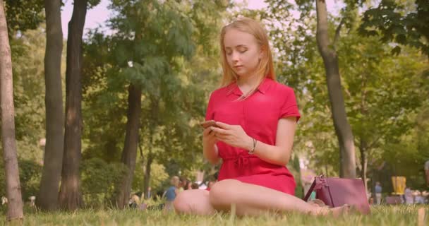 Närbild porträtt av unga blonda långhårig vacker kvinna använda telefonen sitter på gräs i parken utomhus — Stockvideo