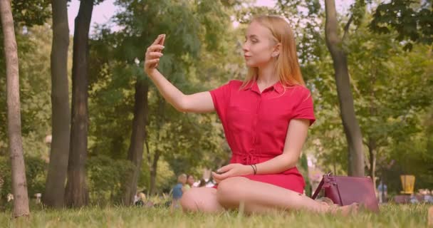 若いブロンドの長い髪の美しい女性のクローズアップ肖像画は、屋外の公園の緑の草の上に座って携帯電話で自分撮りを取ります — ストック動画