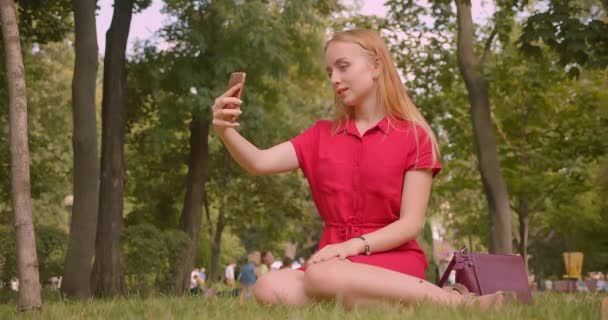 Gros plan portrait de jeune blonde aux cheveux longs belle femelle ayant un appel vidéo au téléphone assis sur l'herbe dans le parc à l'extérieur — Video