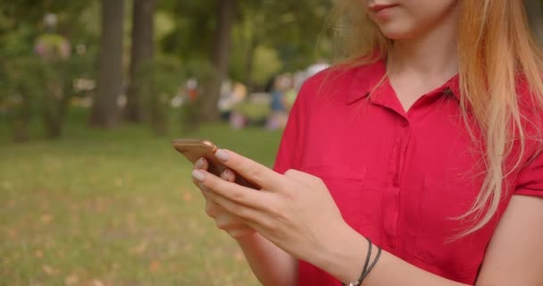 Портрет крупным планом молодой блондинки с длинными волосами симпатичной женщины с помощью телефона, сидящего на траве в парке на открытом воздухе — стоковое видео
