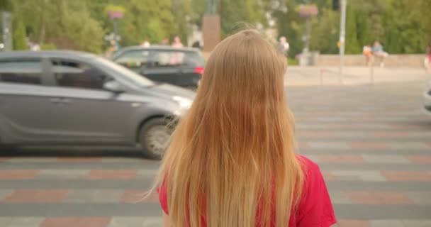 Closeup back view portrait of young blonde long haired beautiful female standing by road with cars passing by outdoors — Stock Video