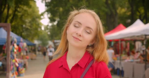 Close-up portret van jonge mooie blanke vrouw in rode jurk kijken naar camera staande in stedelijke stad buitenshuis met markt op achtergrond — Stockvideo