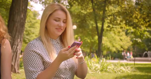 Närbild porträtt av två ganska kvinnliga vänner med hjälp av telefoner som delar roliga inlägg skrattar glatt sitter på filt i parken utomhus — Stockvideo