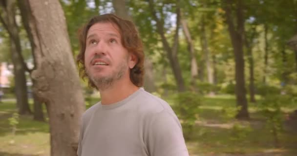 屋外の緑の木と公園に立って幸せな笑顔の白人男性の周りを移動するカメラによるクローズアップポートレート — ストック動画