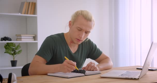 Closeup portrait of young attractive blonde hipster businessman working on laptop taking notes indoors in office — Stock Video