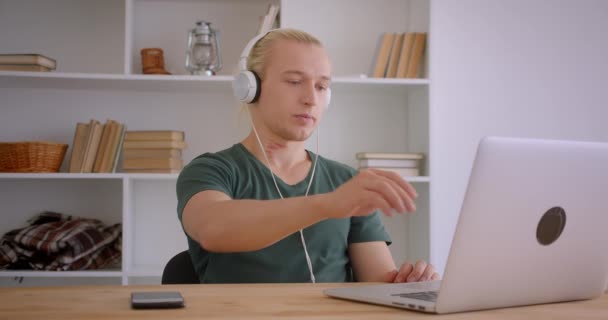 Portrait rapproché de jeune homme d'affaires hipster blond beau dans les écouteurs en utilisant un ordinateur portable à l'intérieur dans le bureau avec des étagères sur fond — Video
