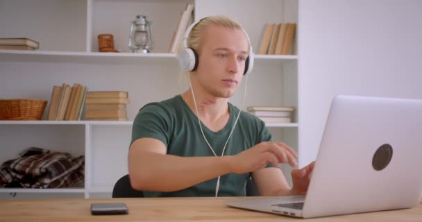 Closeup portré fiatal csinos szőke csípő üzletember fejhallgató segítségével laptop, hogy nyugodt beltéri hivatalban — Stock videók
