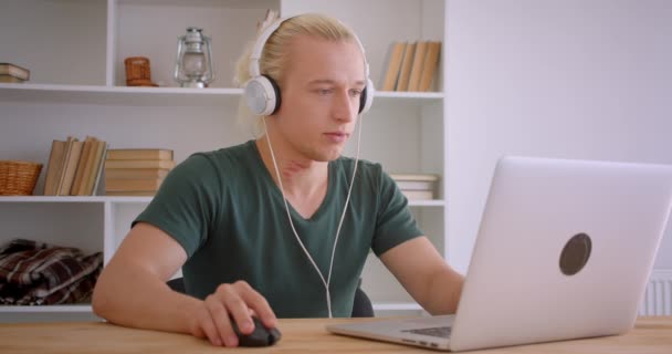 Close-up retrato de jovem bonito loira hipster empresário em fones de ouvido usando laptop ouvindo música dentro de casa no escritório — Vídeo de Stock