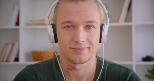 Zbliżenie portret młodego atrakcyjny blond Hipster człowiek w słuchawkach patrząc na aparat uśmiechnięty szczęśliwie w pomieszczeniu — Wideo stockowe