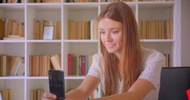 Genç güzel beyaz iş kadını kapalı kütüphane dizüstü önünde oturan telefonda görüntülü arama olan Closeup portre