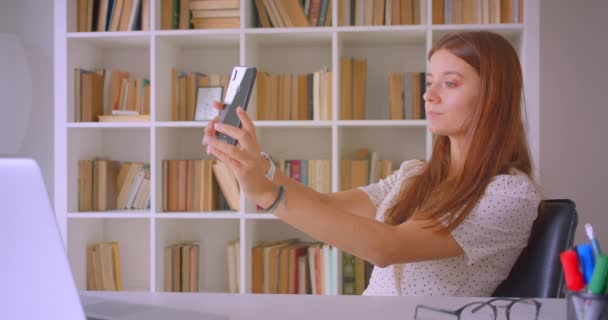 Zbliżenie Portret młodej kaukaski bizneswoman biorąc selfie na telefon siedzi przed laptopem w bibliotece w pomieszczeniu — Wideo stockowe