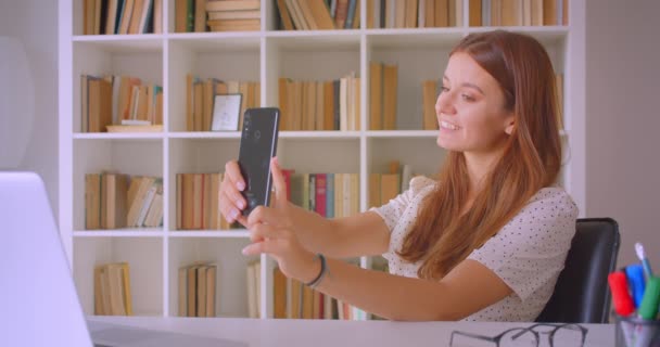 Closeup portré fiatal kaukázusi üzletasszony figyelembe selfies telefonon mosolyogva boldogan ül előtt laptop könyvtárban beltéri — Stock videók