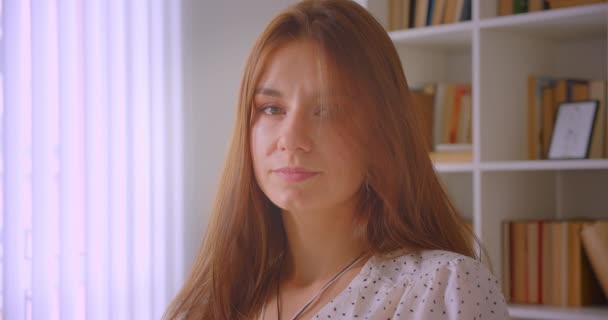 Close-up portret van jonge Kaukasische zakenvrouw kijken naar camera in bibliotheek binnenshuis met boekenkasten op achtergrond — Stockvideo