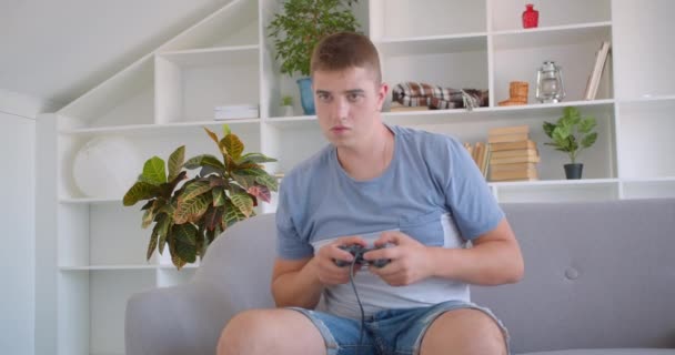 Gros portrait de l'homme caucasien attrayant adulte jouant à des jeux vidéo en utilisant la console de jeu étant nerveux assis sur le canapé à l'intérieur — Video