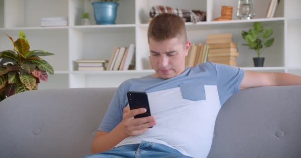 Крупним планом портрет дорослого привабливого білоруського чоловіка, який переглядає по телефону, сидячи на дивані в приміщенні — стокове відео