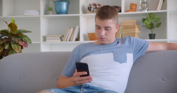 Nahaufnahme Porträt eines erwachsenen attraktiven kaukasischen Mannes, der mit seinem Handy aufgeregt auf der Couch im Haus sitzt — Stockvideo