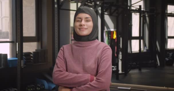 Крупный план портрета молодой привлекательной спортивной мусульманки, смотрящей в камеру со скрещенными руками на груди, весело улыбающейся в спортзале в помещении — стоковое видео