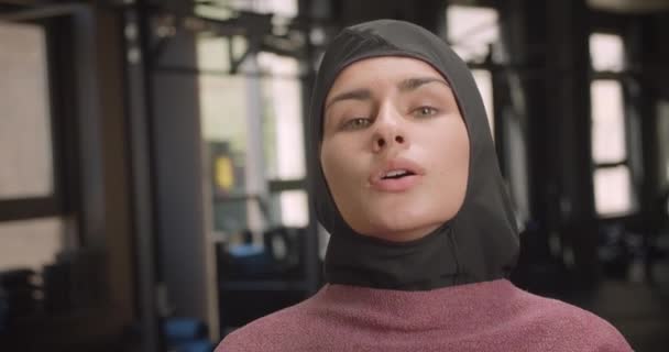 Крупный план портрета молодой привлекательной спортивной мусульманки, смотрящей в камеру, мотивированной стоящей в спортзале в помещении — стоковое видео