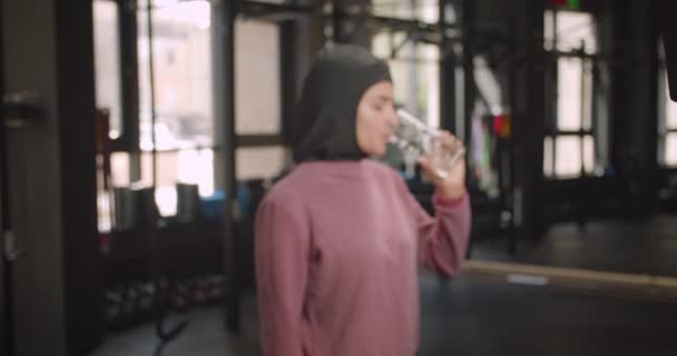 Primer plano retrato de joven atractivo atlético musulmán hembra mirando la cámara de agua potable en el gimnasio en interiores — Vídeo de stock