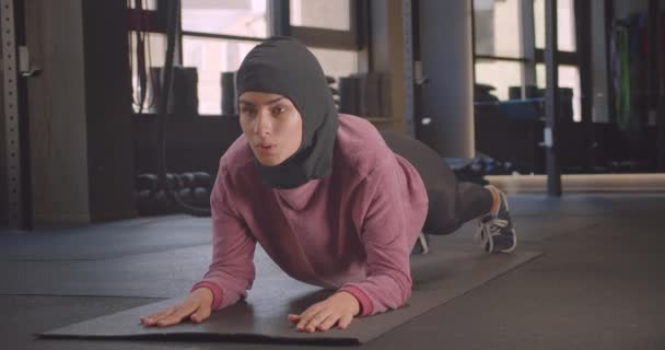 Крупним планом портрет молодої визначеної атлетичної мусульманки в хіджабі роблять тренувальні дошки в тренажерному залі в приміщенні — стокове відео
