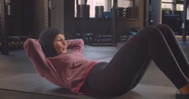 Close-up portret van jonge vastberaden atletische moslim vrouw in hijab doen ABS oefening trainen in de sportschool binnenshuis — Stockvideo