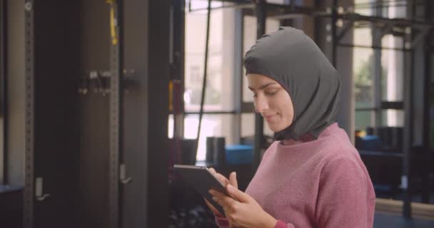 Retrato de primer plano de una joven atlética musulmana en hijab usando una tableta mirando a la cámara sonriendo felizmente en el gimnasio interior — Vídeo de stock