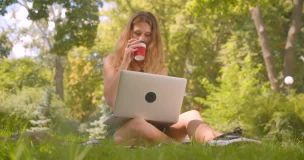 Крупним планом портрет молодої досить кавказької жінки з використанням ноутбука п'є каву, що сидить на килимі на траві в парку — стокове відео