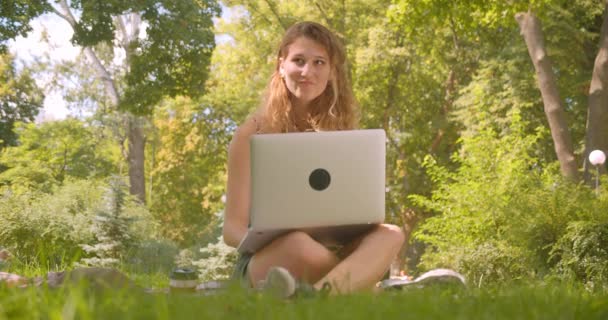 Närbild porträtt av unga ganska kaukasiska kvinna med hjälp av laptop leende lyckligt sittande på mattan på gräs i parken — Stockvideo
