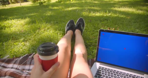 Primer plano retrato de joven bastante caucásico piernas femeninas niña sentada en la alfombra sosteniendo el café con el ordenador portátil con pantalla azul en el parque al aire libre — Vídeos de Stock