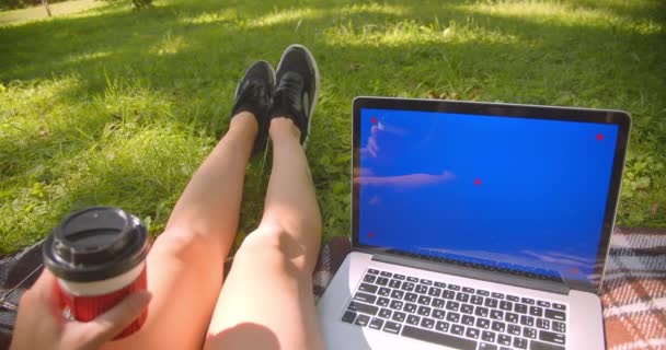 Nahaufnahme Porträt der jungen hübschen kaukasischen weiblichen Beine Mädchen sitzt auf Teppich hält Kaffee mit Laptop mit blauem Chroma-Bildschirm im Park im Freien — Stockvideo