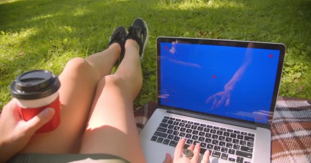 Primer plano retrato de joven bastante caucásico piernas femeninas niña sentada en la alfombra sosteniendo el café con el ordenador portátil con pantalla de croma azul clave en el parque al aire libre — Vídeos de Stock