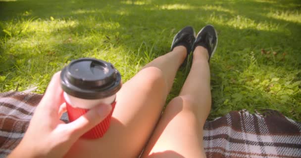 Closeup portrait of young pretty caucasian female legs girl sitting on rug holding coffee in park outdoors — Stock Video