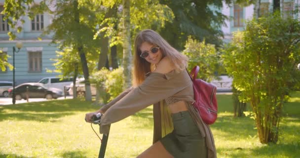 Primer plano retrato de joven guapa caucásica hembra en gafas de sol de pie con patinete scooter en el parque — Vídeo de stock