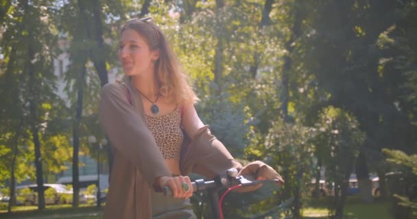 Retrato de close-up de jovem muito caucasiano fêmea de pé com pontapé scooter olhando para a câmera no parque — Vídeo de Stock